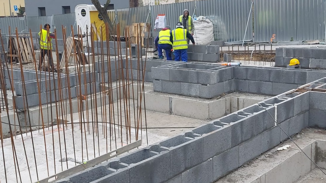 Wznoszenie fundamentów z szarych bloczków betonowych na placu budowy, widoczni pracownicy w żółtych kamizelkach, zbrojenie pionowe i poziomica murarska w kadrze.