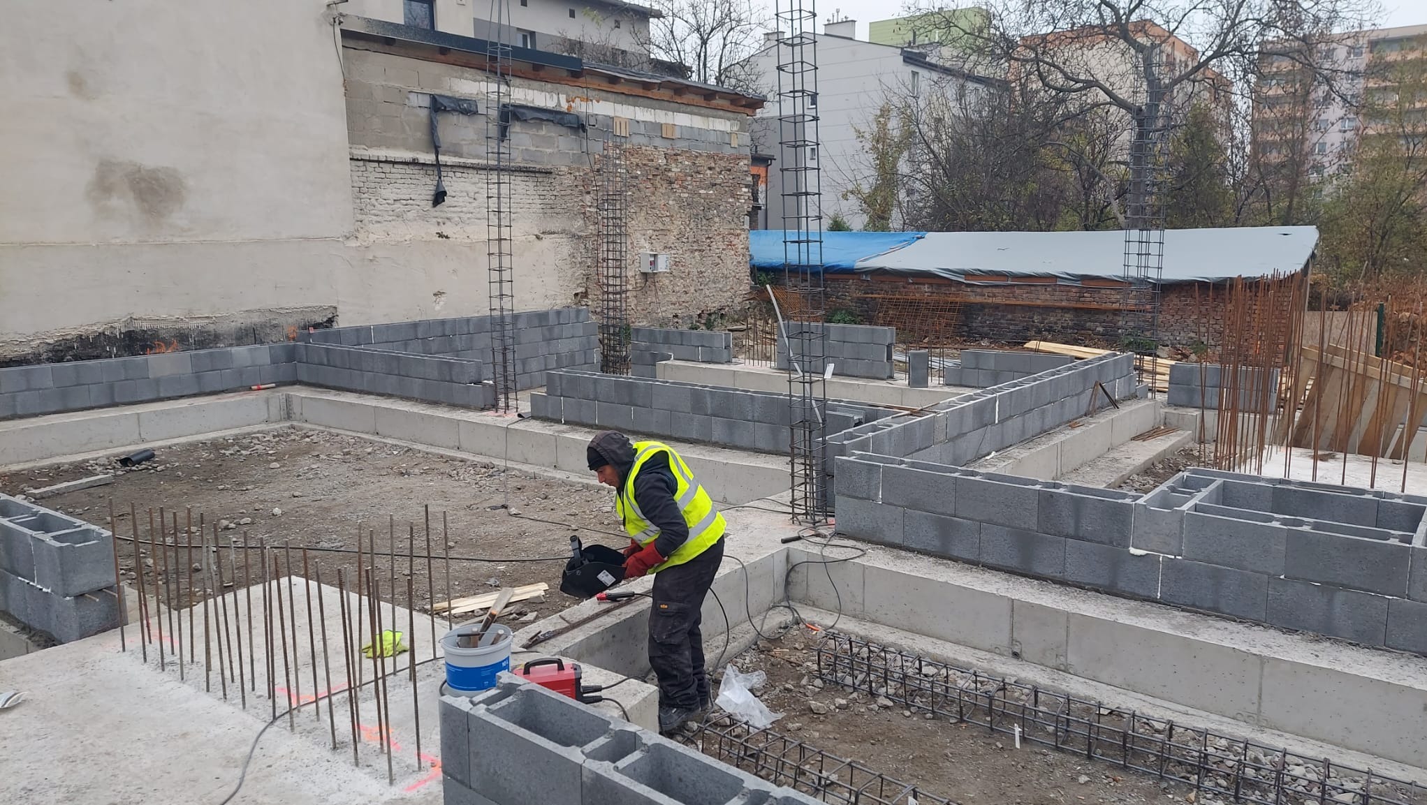 Pracownik w kamizelce odblaskowej tnie bloczki betonowe na placu budowy fundamentów domu, widoczne zbrojenia i częściowo wymurowane ściany fundamentowe.