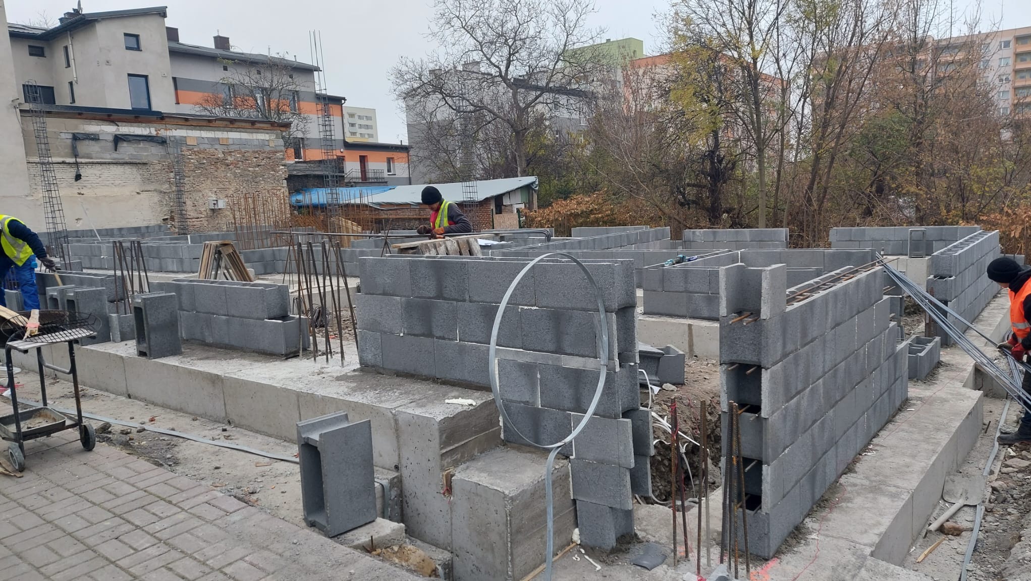 Wznoszenie fundamentów i ścian parteru z bloczków betonowych na placu budowy, widoczne zbrojenia i pracownicy w odblaskowych kamizelkach.