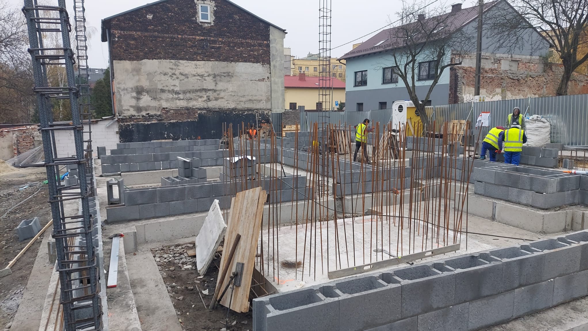 Fundamenty domu z bloczków betonowych na placu budowy, widoczne zbrojenia i pracownicy w odblaskowych kamizelkach.