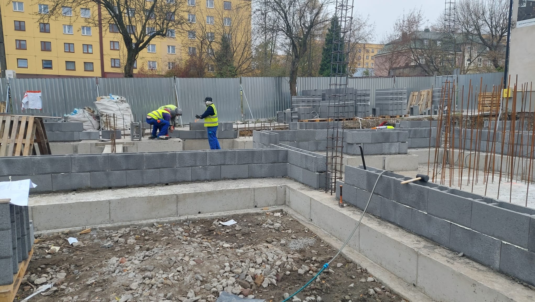 Wznoszenie fundamentów z bloczków betonowych na placu budowy; widoczni pracownicy w odblaskowych kamizelkach, zbrojenie, palety z bloczkami oraz budynki w tle.