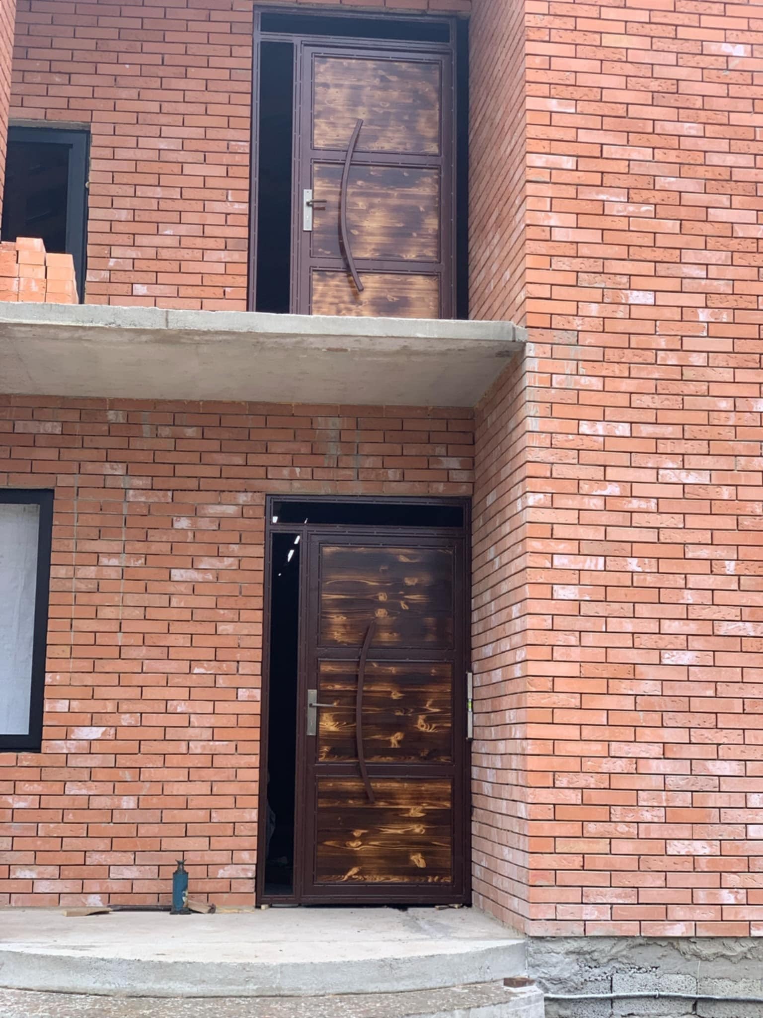 Wznoszenie dwukondygnacyjnego domu z czerwonej cegły z zamontowanymi, brązowymi, metalowymi drzwiami wejściowymi na parterze i pierwszym piętrze.