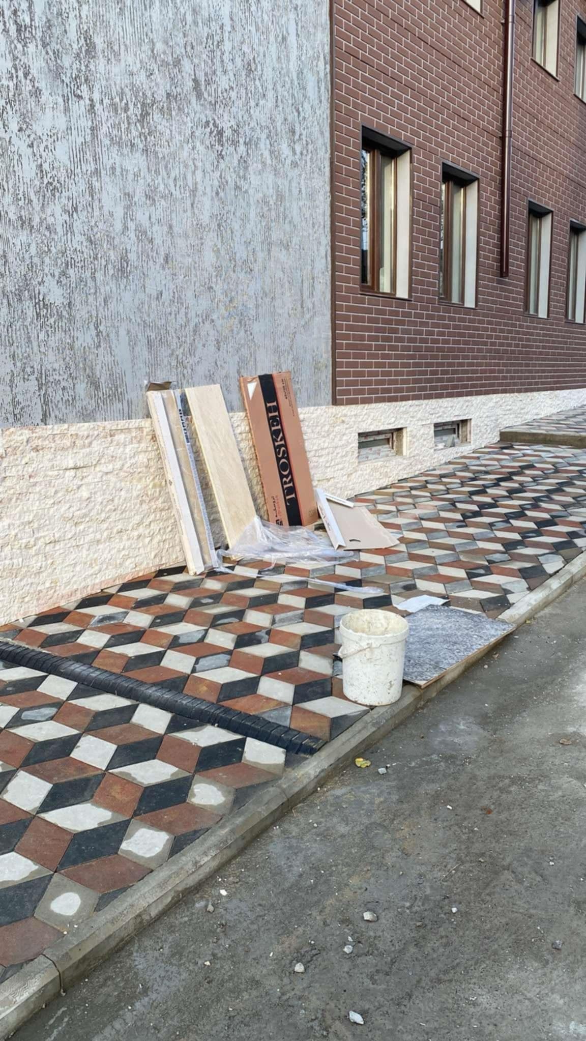 Ułożona kostka brukowa w geometryczny wzór 3D przed budynkiem z cegły, widoczne paczki kostki i wiadro.