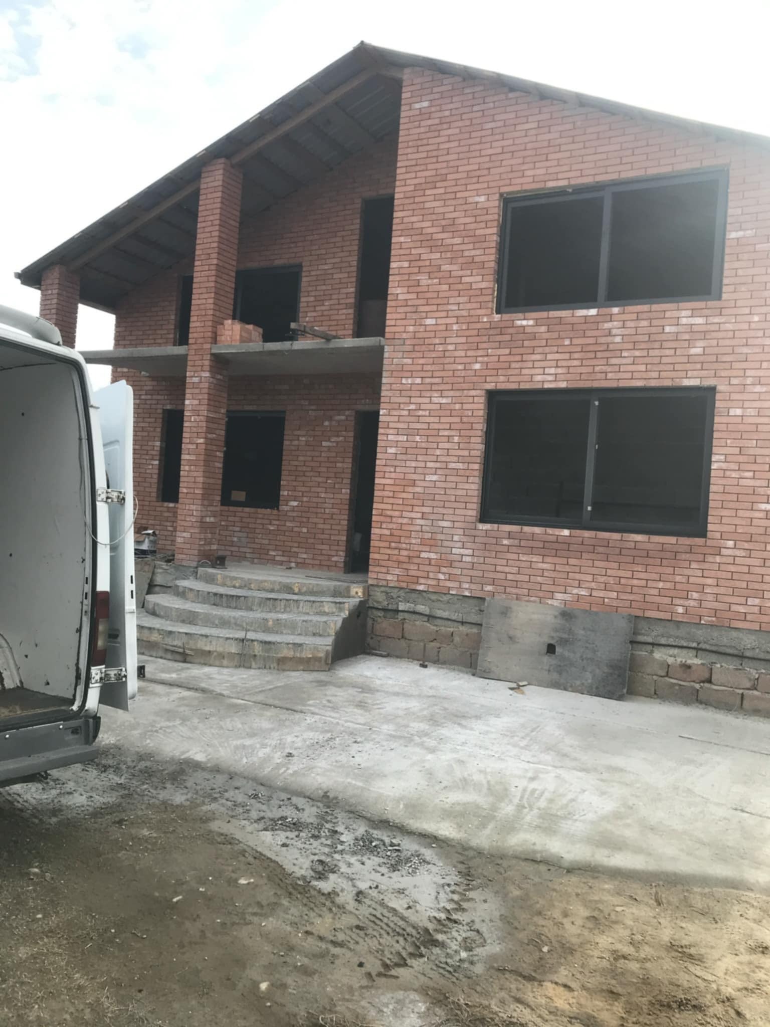 Surowy stan dwupiętrowego domu z czerwonej cegły z czarnymi ramami okiennymi, widok z zewnątrz, z betonowymi schodami wejściowymi i częściowo widocznym białym vanem po lewej stronie.