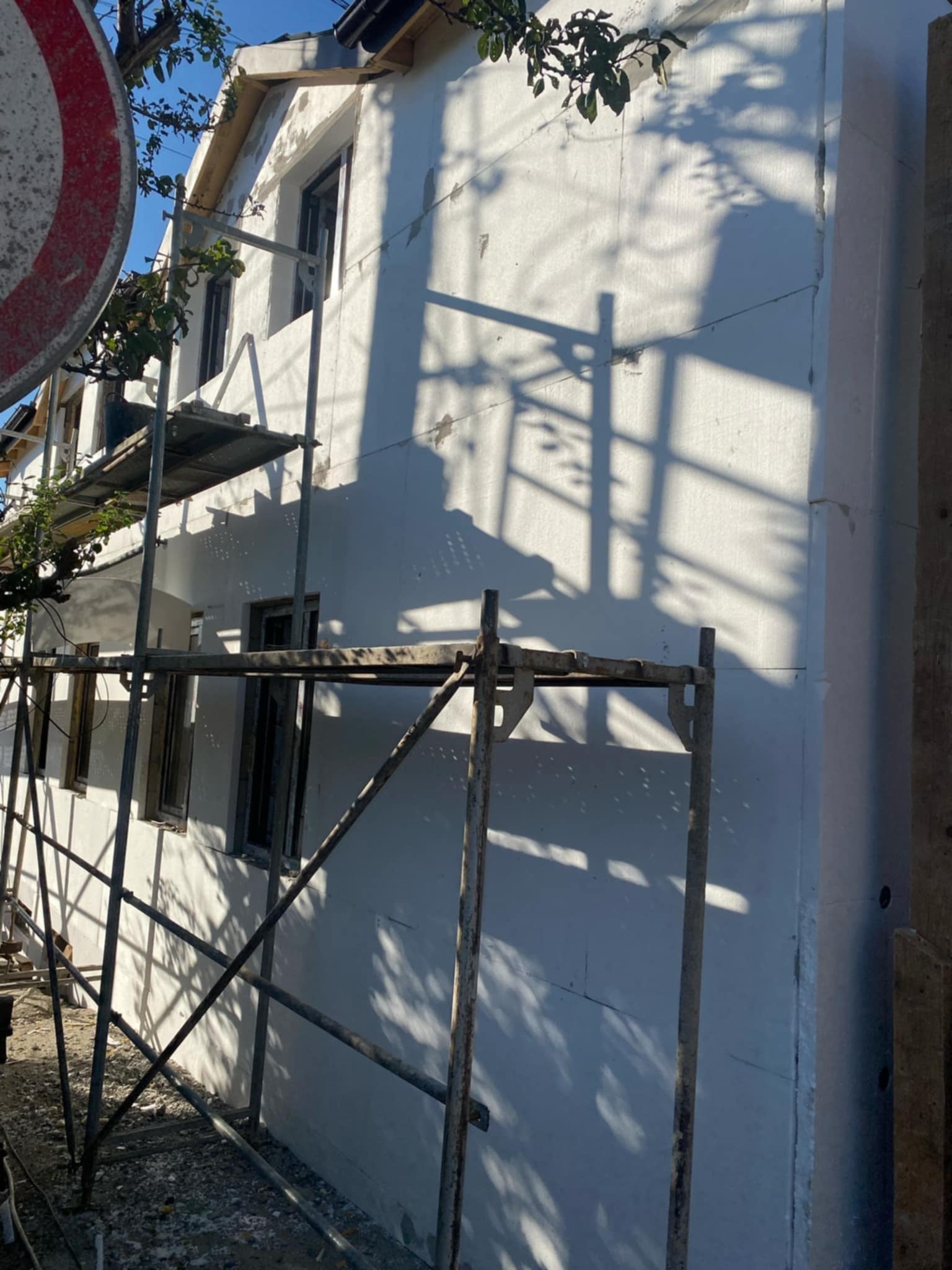 Ściana budynku w trakcie ocieplania, widoczne rusztowanie i cień rzucany przez gałęzie drzewa na elewacji.