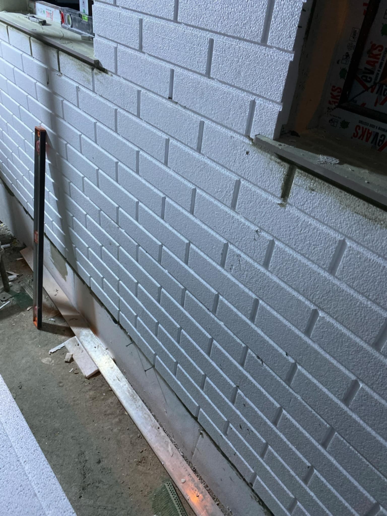 Biała ściana z imitacją cegły, wykonana z paneli styropianowych, z widoczną poziomicą i oknem w tle, w trakcie montażu.