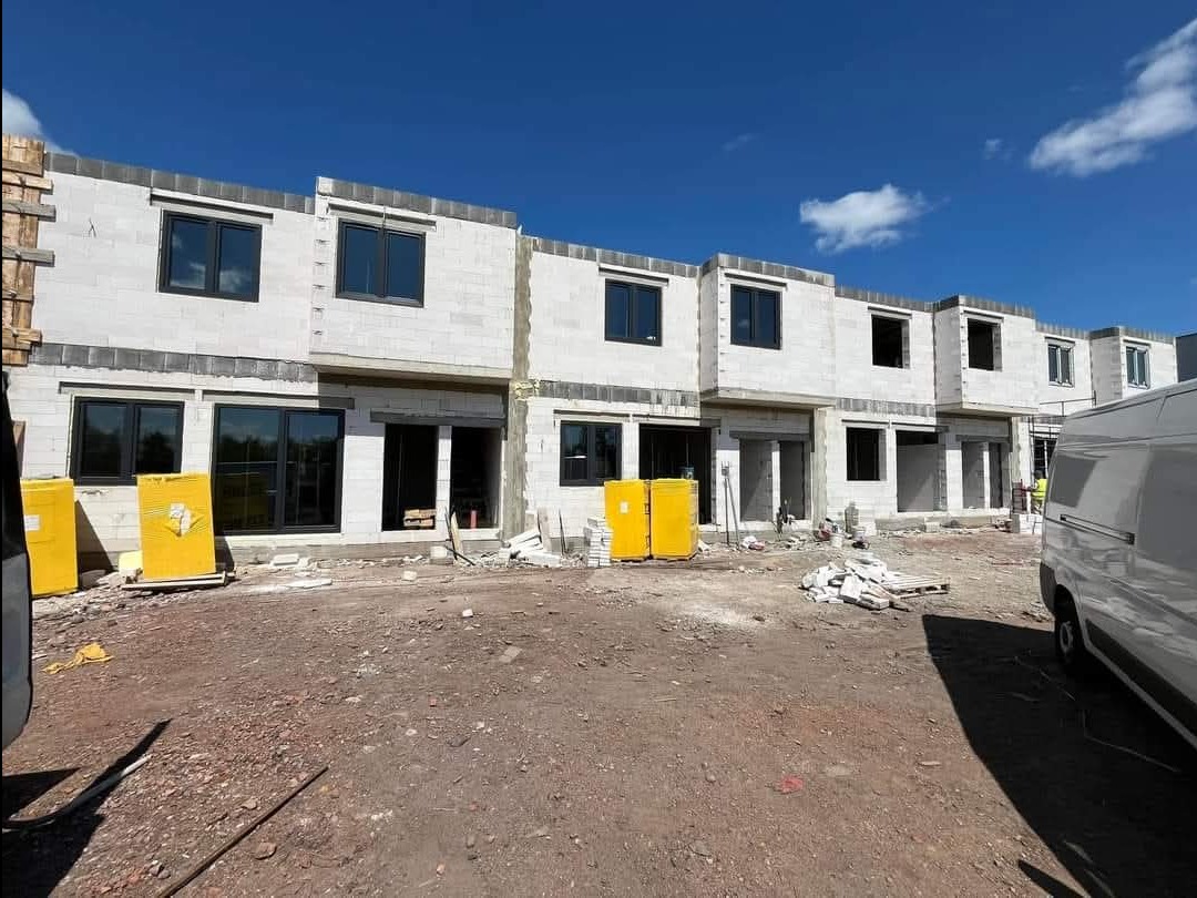 Wznoszenie szeregu domów z betonu komórkowego, widoczne pakiety żółtej izolacji termicznej i biały van w tle, błękitne niebo.