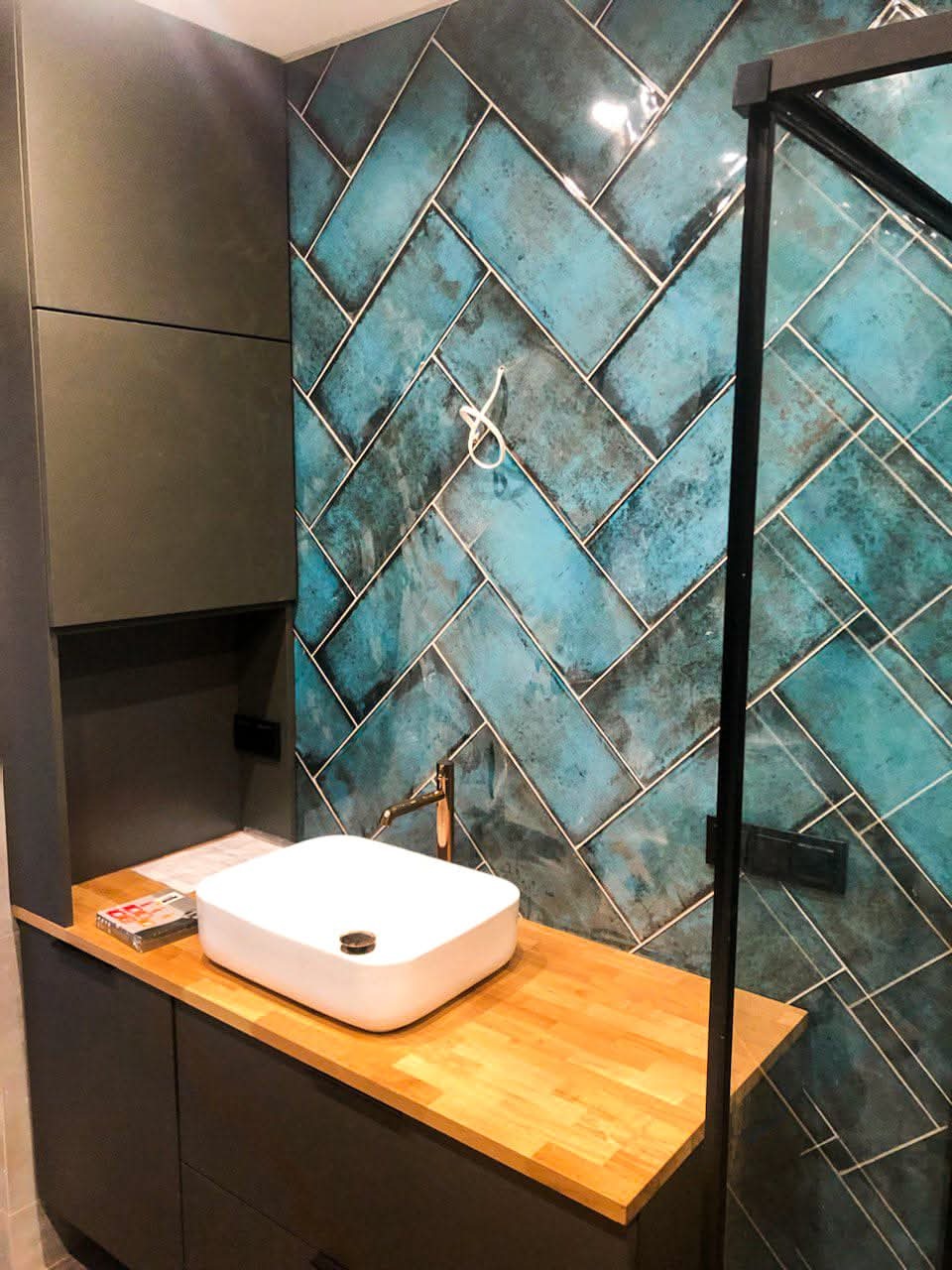 Nowoczesna łazienka z umywalką nablatową i turkusową ścianą z płytek ułożonych w jodełkę. Widoczna kabina prysznicowa z czarnymi profilami i drewniany blat.