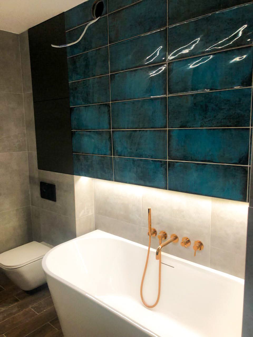 Nowoczesna łazienka z wolnostojącą wanną, baterią w kolorze miedzi i turkusowymi płytkami na ścianie. Podświetlenie LED nad wanną. Widoczna toaleta i drewnopodobna podłoga.