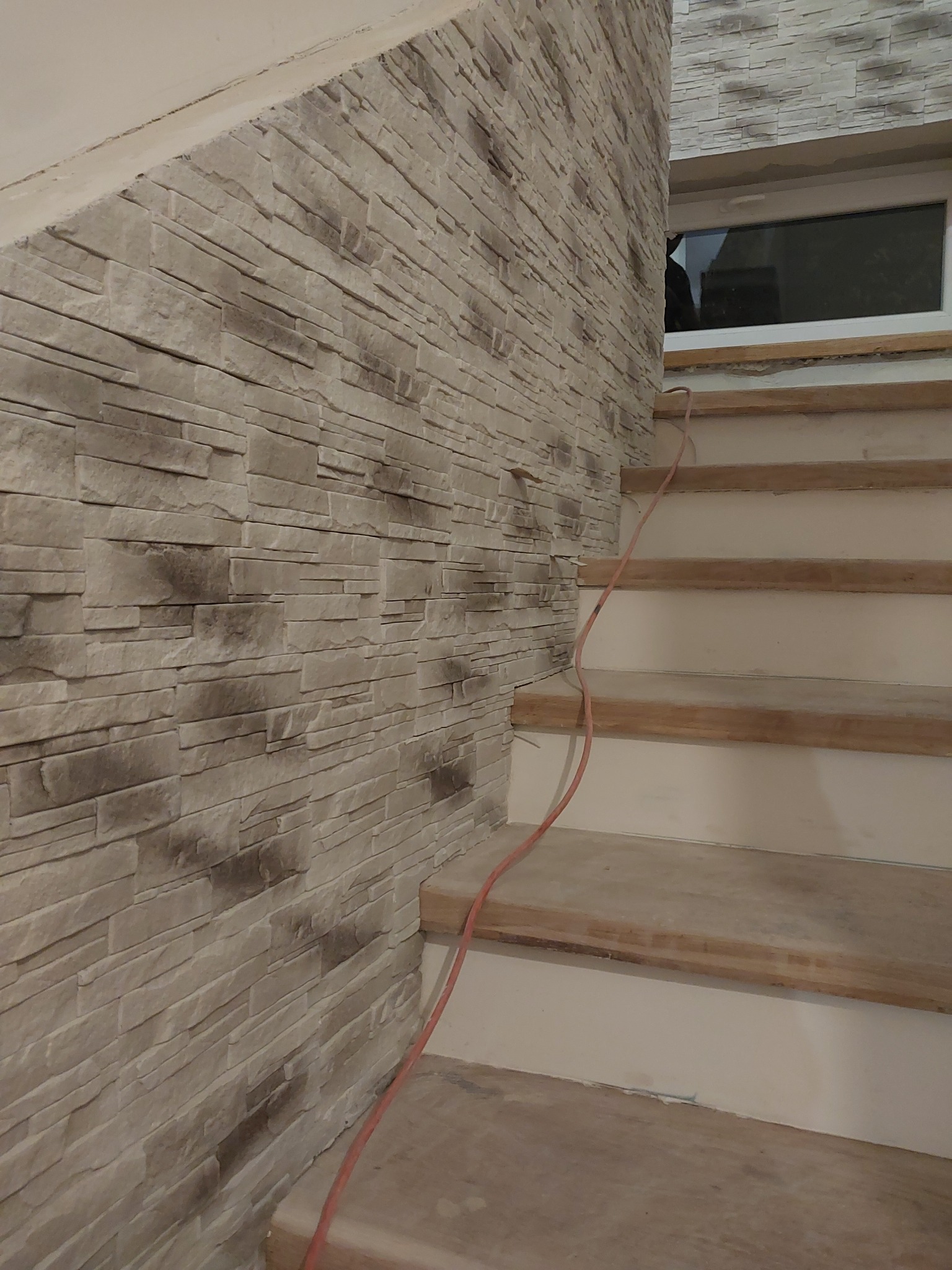 Ściana wykończona dekoracyjną okładziną kamienną w odcieniach beżu, sąsiadująca z drewnianymi schodami w trakcie montażu. Widoczne okno w tle.