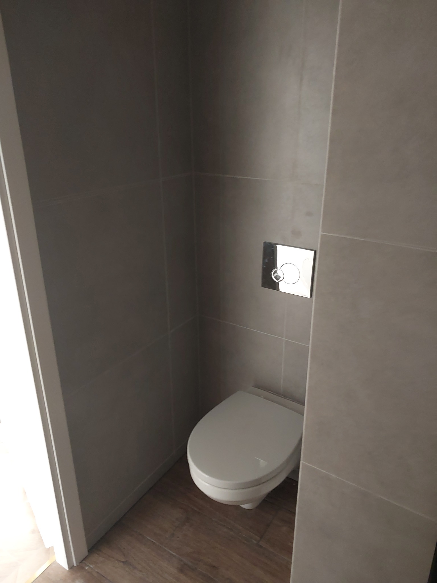 Nowoczesna toaleta podwieszana z chromowanym przyciskiem spłukującym, wykończona szarymi płytkami i drewnopodobną podłogą. Minimalistyczny design.