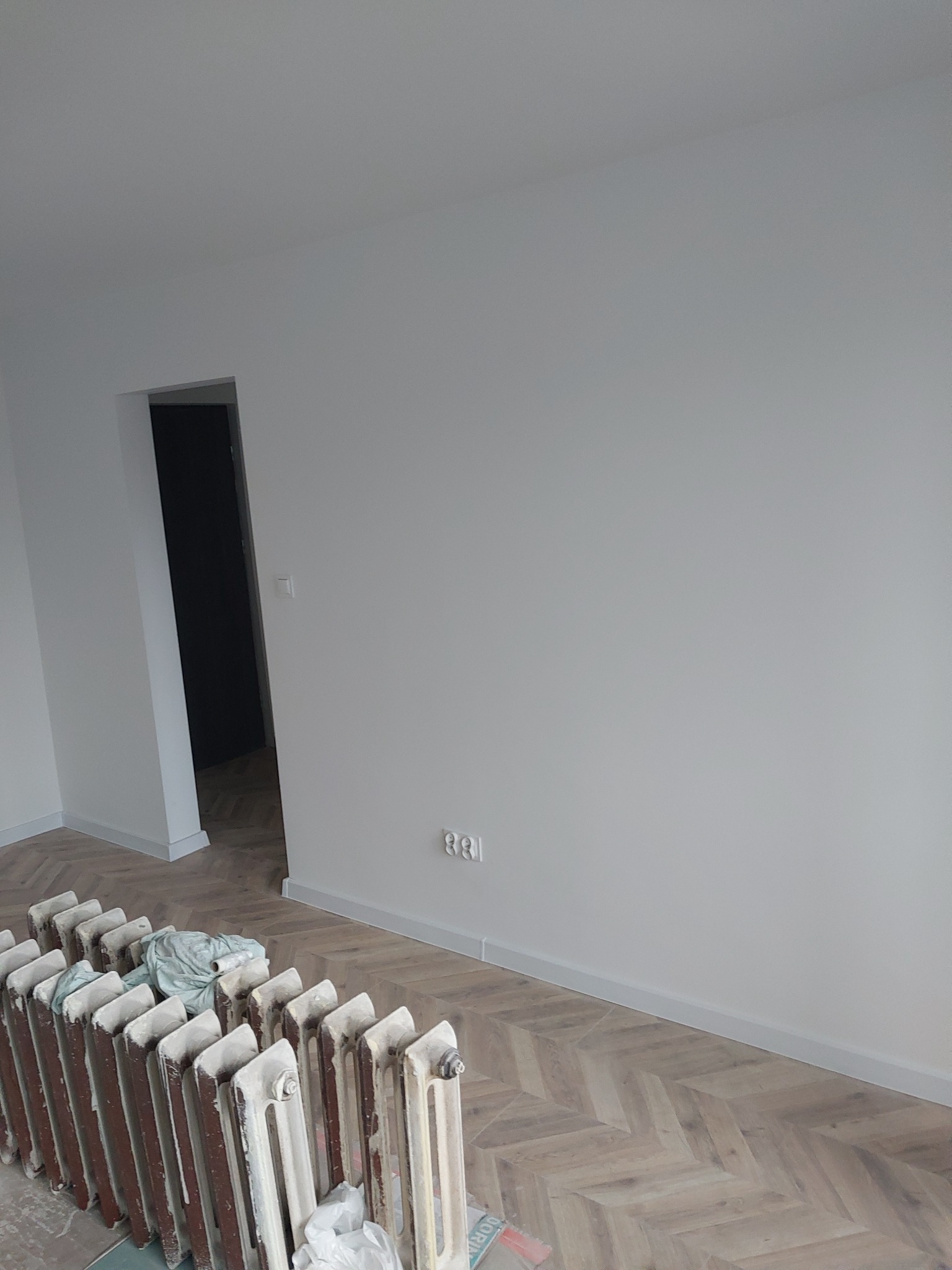 Białe ściany po malowaniu, parkiet w jodełkę, grzejnik żeliwny w trakcie renowacji. Widoczne ślady farby i zabezpieczenia podłogi. W tle otwarte drzwi.