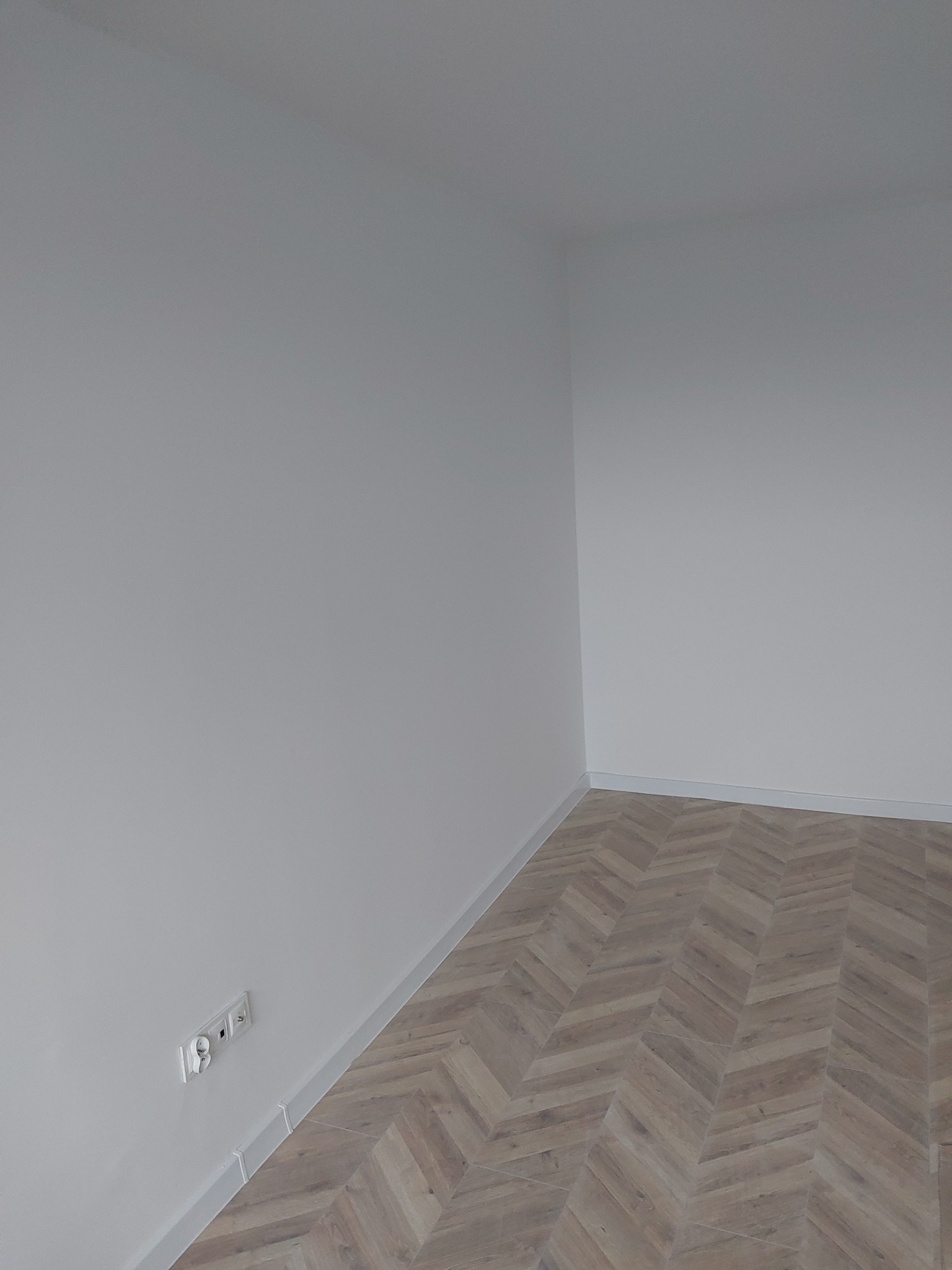 Jasne panele ułożone w jodełkę w rogu pokoju z białymi ścianami i listwami przypodłogowymi. Widoczne gniazdka elektryczne. Minimalistyczne wykończenie wnętrza.