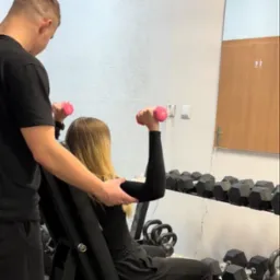 Kobieta ćwiczy unoszenie hantli pod nadzorem mężczyzny w siłowni, widoczny stojak z hantlami i drzwi z oznaczeniem toalety.