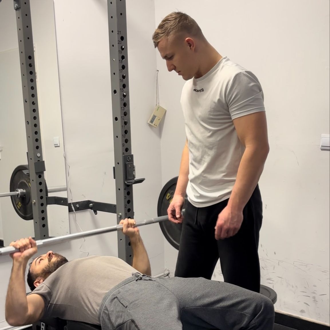 Mężczyzna ćwiczy wyciskanie sztangi leżąc na ławce, a drugi mężczyzna w białej koszulce go asekuruje i obserwuje w siłowni.