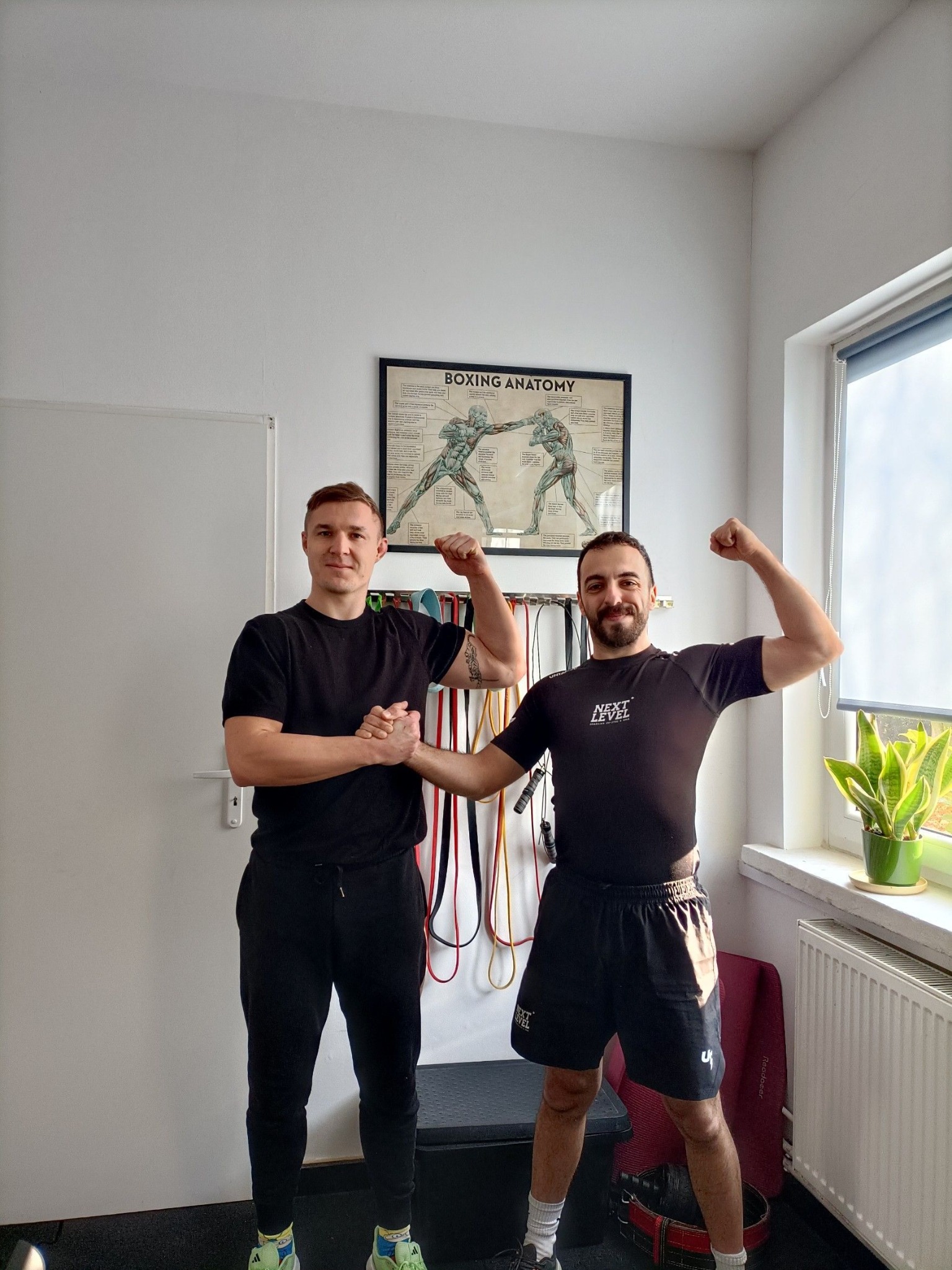 Dwóch mężczyzn w sportowych strojach, jeden trzyma drugiego za rękę, obaj prezentują napięte bicepsy na tle plakatu z anatomią boksu i wiszących gum do ćwiczeń.