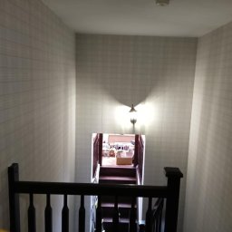 Usługi Remontowe Stanisław Gawronowicz - Klatka schodowa w domu z tapetą w kratkę, widok z góry na schody prowadzące w dół, oświetlone lampą ścienną.