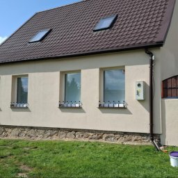 Usługi Remontowe Stanisław Gawronowicz - Beżowy dom jednorodzinny z brązowym dachem, widoczne okna z kratami, adres na ogrodzeniu, drabina oparta o ścianę i wiadro z farbą na trawie.