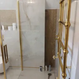 Nowoczesna kabina prysznicowa z elementami w kolorze złotym, widoczny grzejnik łazienkowy w tym samym kolorze, marmurowe płytki na ścianach i podłodze, oraz drobne akcesoria łazienkowe.