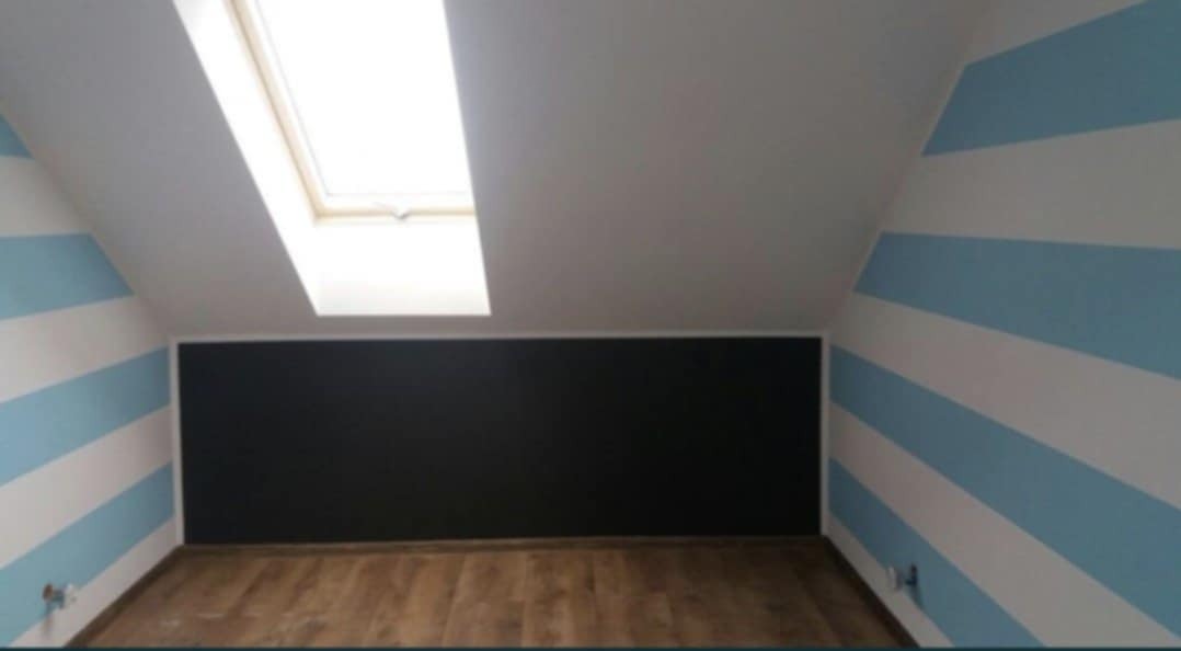 Pokój na poddaszu z pomalowanymi ścianami w biało-błękitne pasy i czarną ścianą tablicową, oświetlony przez okno dachowe.