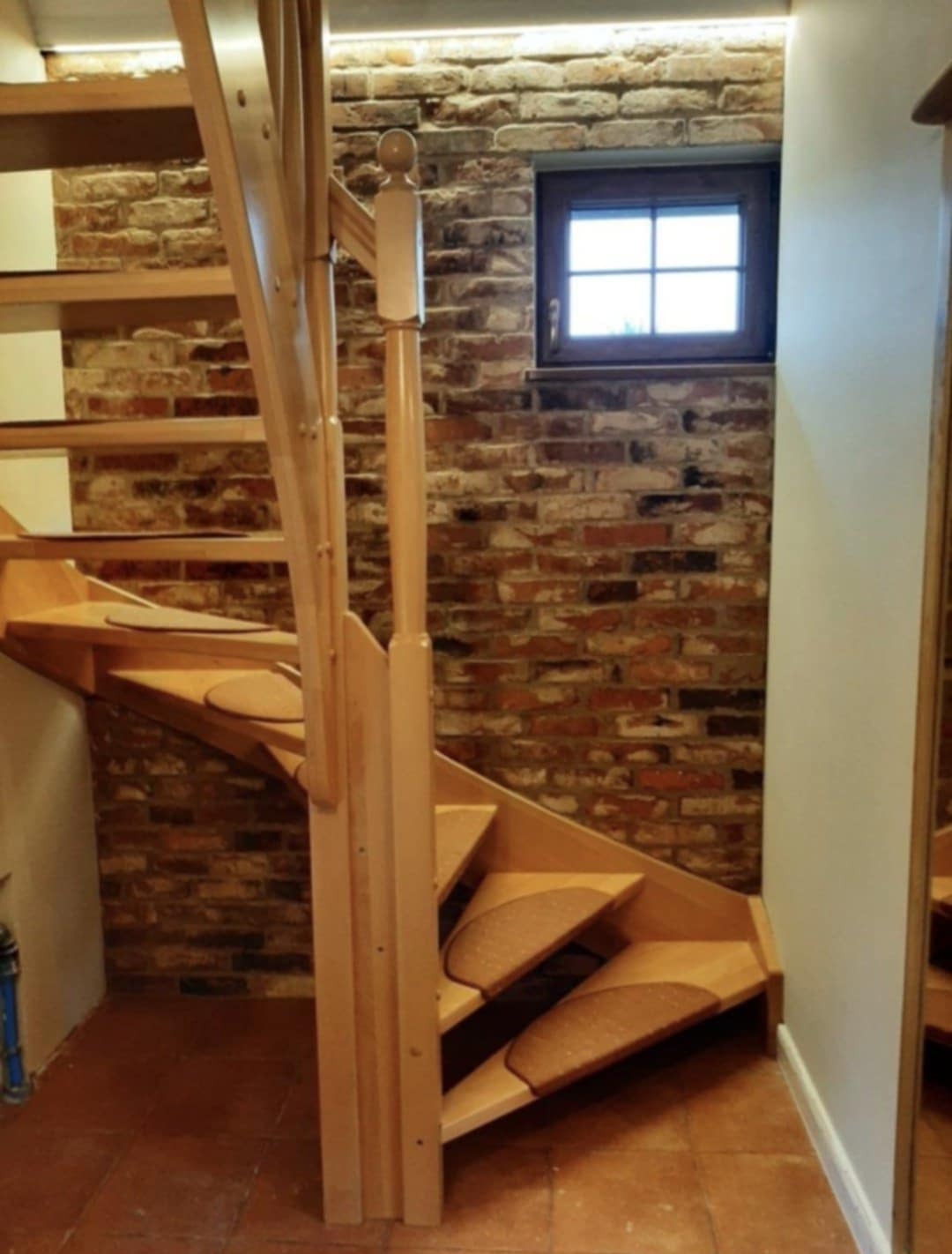 Drewniane schody spiralne z brązowymi nakładkami na stopniach, ustawione przy ceglanej ścianie z małym oknem.