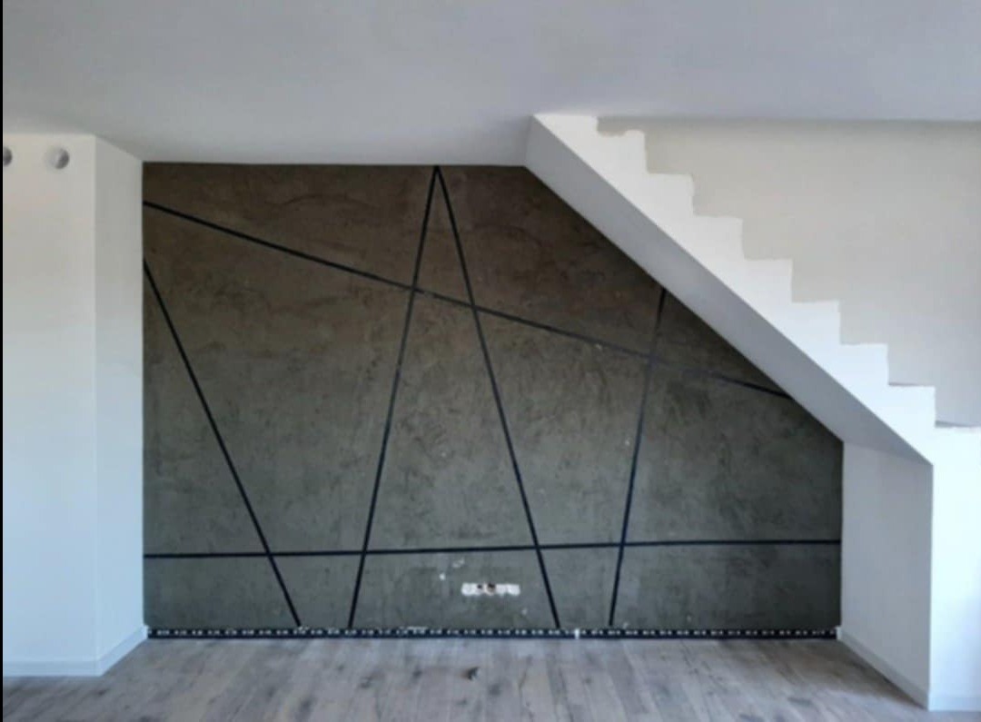 Ściana z dekoracyjną strukturą i geometrycznym wzorem z czarnych linii, białe schody w tle, jasna podłoga.