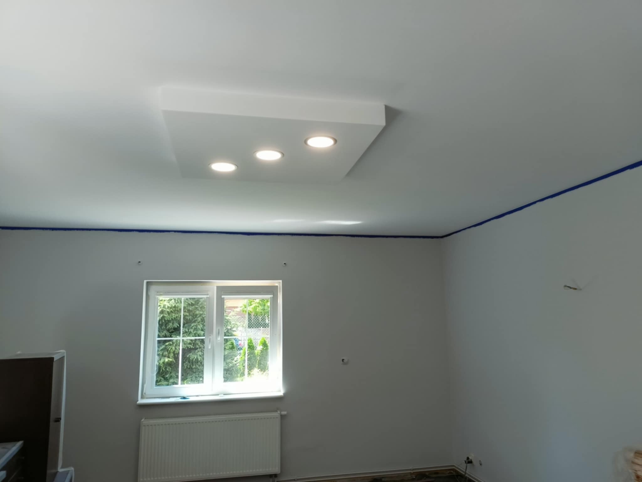 Wnętrze pokoju po odświeżeniu, z widocznym sufitem podwieszanym z trzema punktami świetlnymi, ściany pomalowane na biało, zabezpieczone niebieską taśmą malarską przy suficie, okno z widokiem...