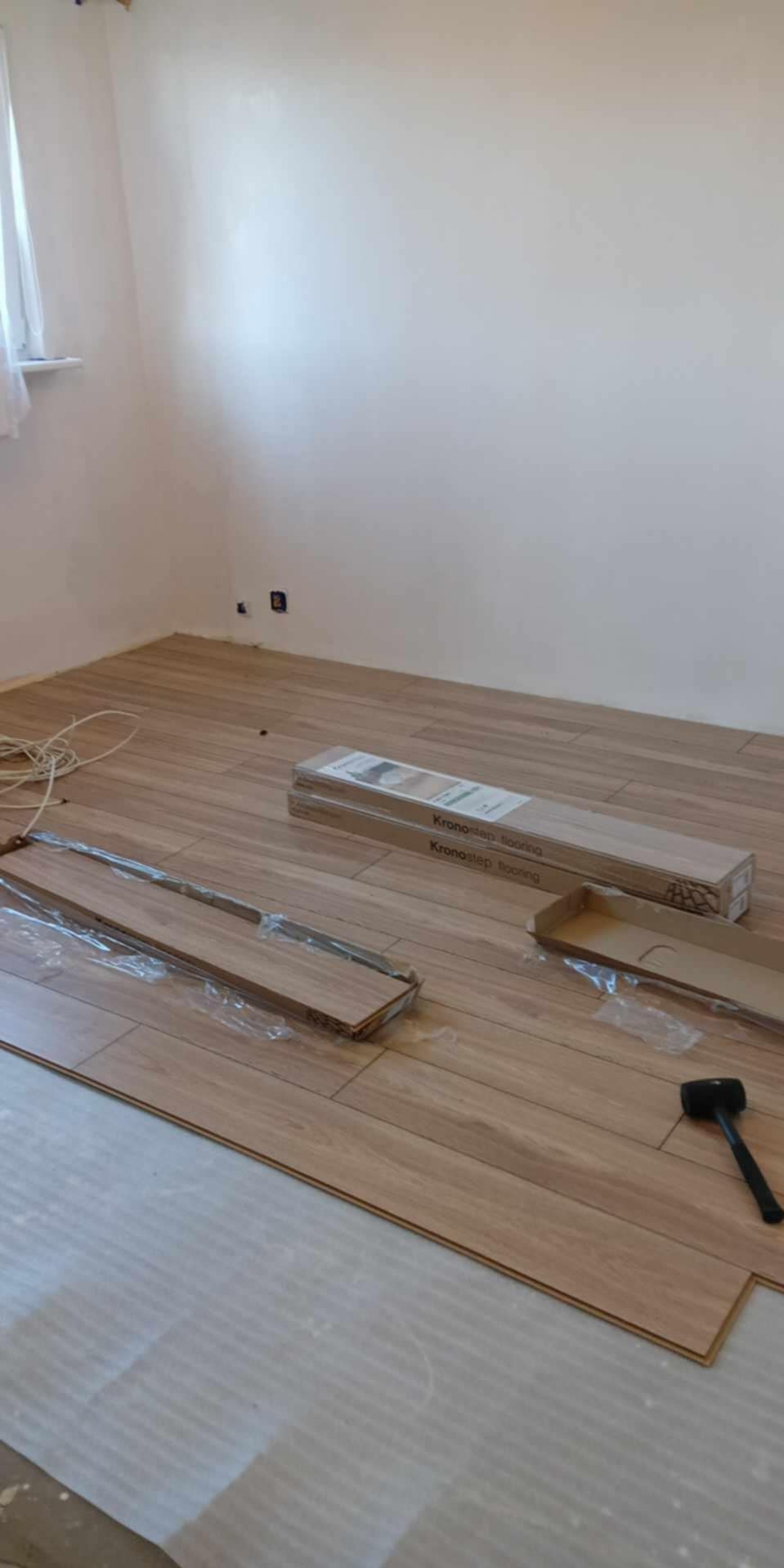 Układanie paneli podłogowych, widoczne paczki paneli Kronostep, młotek gumowy i fragment podkładu pod panele.