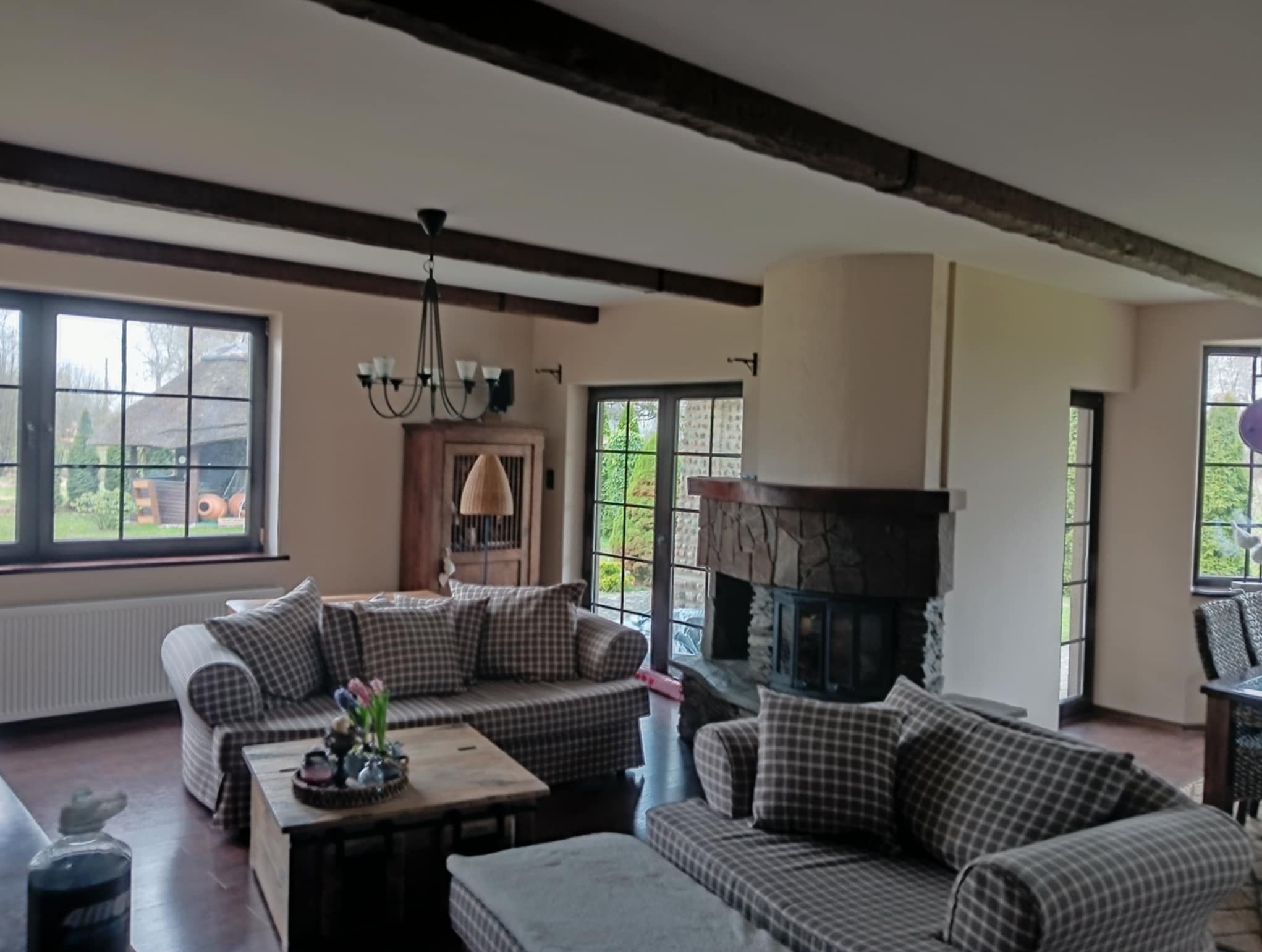 Jasne wnętrze salonu z beżowymi ścianami, drewnianymi belkami stropowymi, kamiennym kominkiem i sofami w kratę, widok na ogród przez okna.