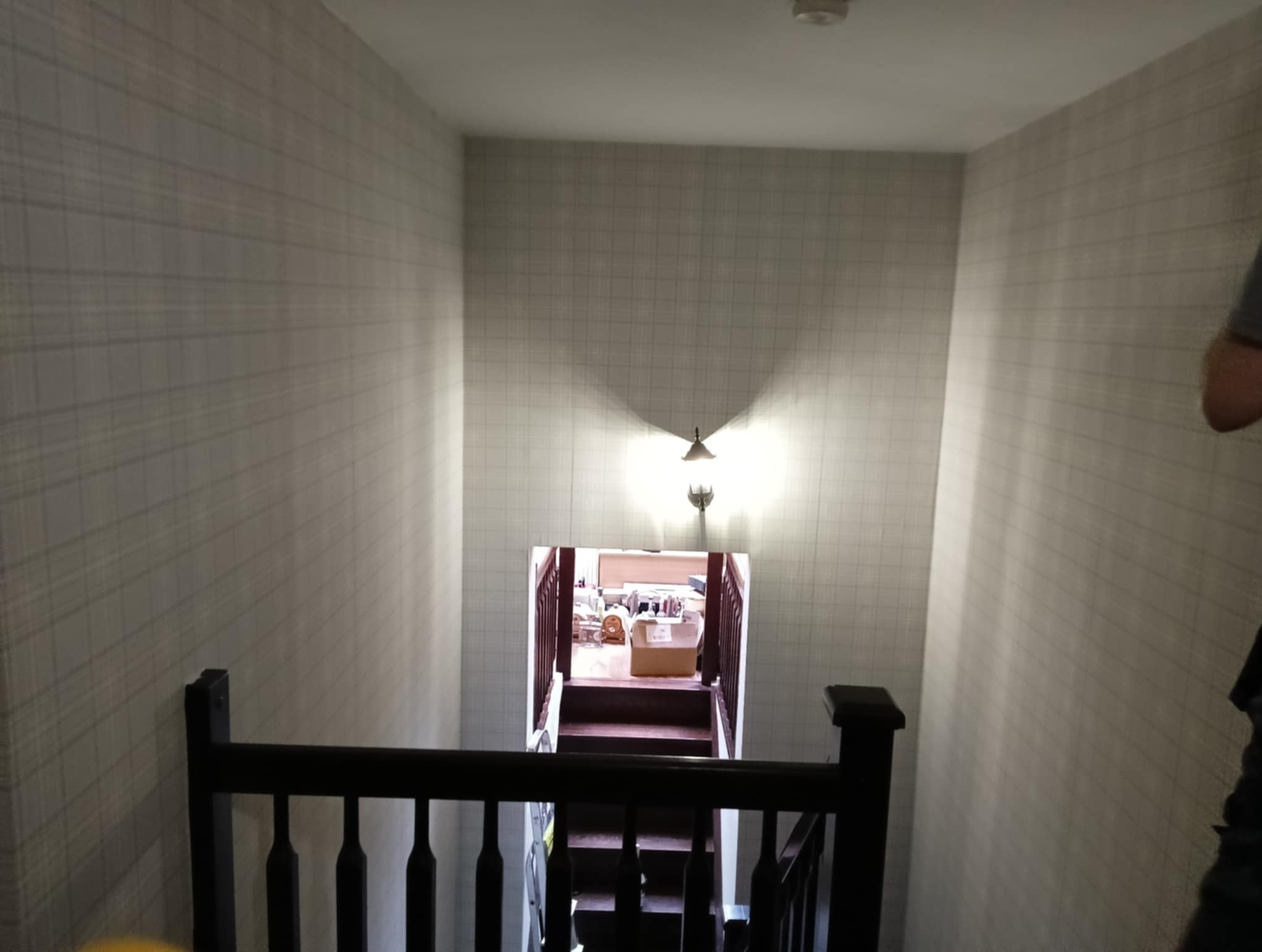 Klatka schodowa w domu z tapetą w kratkę, widok z góry na schody prowadzące w dół, oświetlone lampą ścienną.