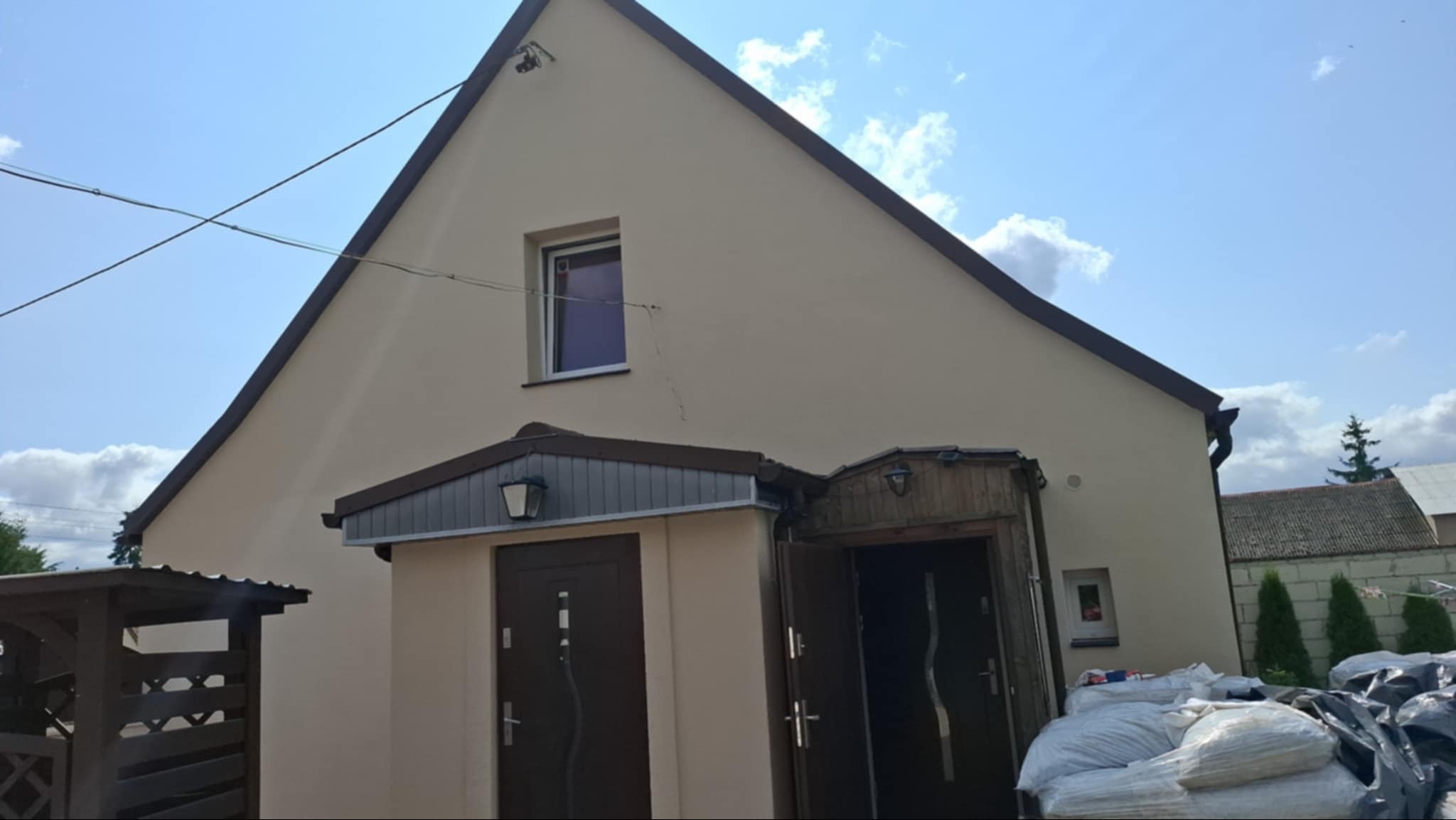 Beżowy dom jednorodzinny z dwoma wejściami, widoczne worki z materiałem budowlanym przed jednym z wejść, na elewacji widoczna pionowa rysa pod oknem.