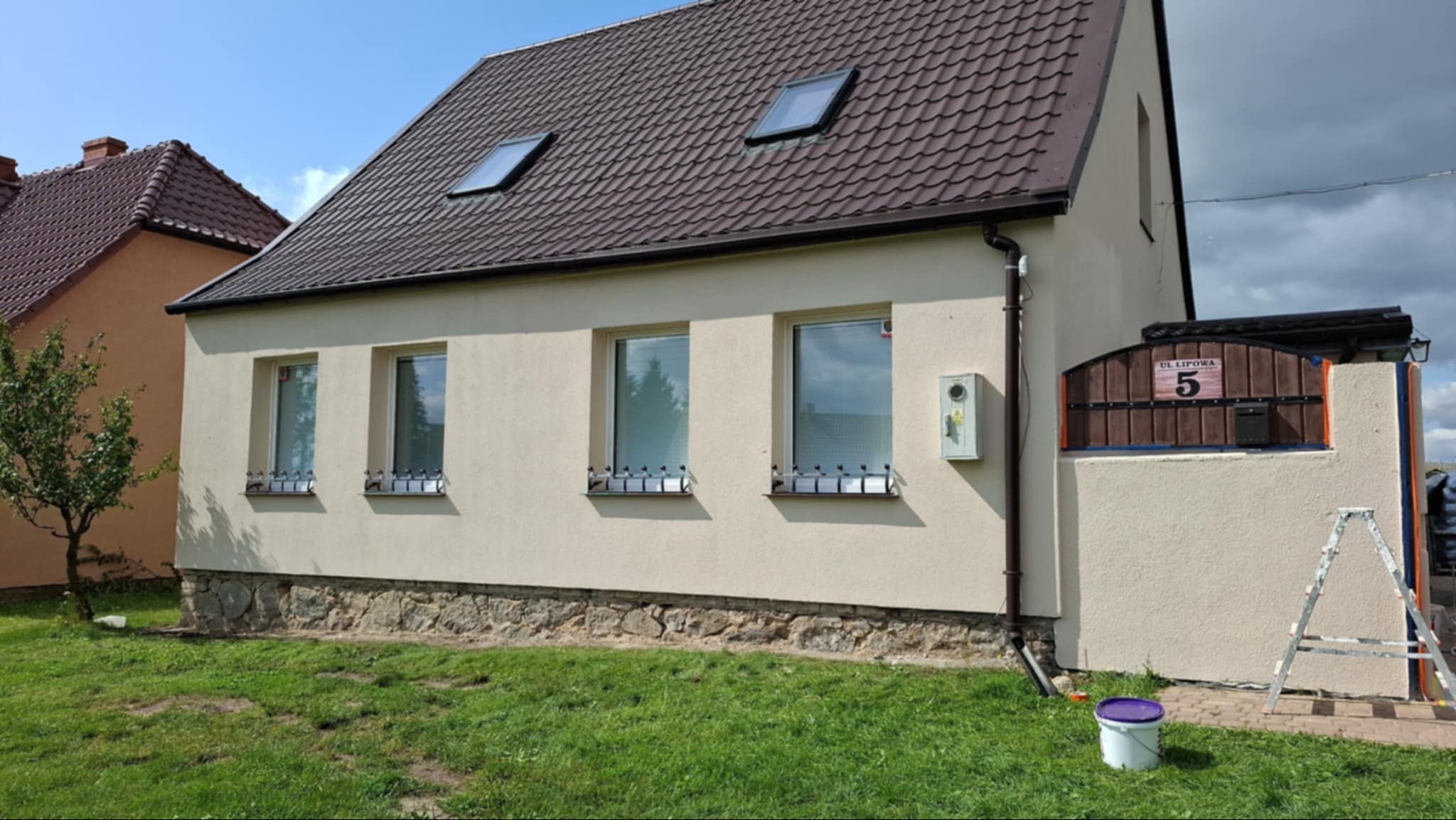 Beżowy dom jednorodzinny z brązowym dachem, widoczne okna z kratami, adres na ogrodzeniu, drabina oparta o ścianę i wiadro z farbą na trawie.