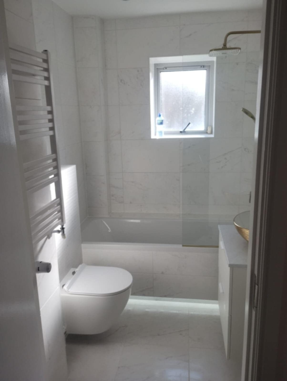 Nowoczesna łazienka z białymi marmurkowymi płytkami, podświetlaną wanną, wiszącą toaletą i złotą umywalką nablatową.