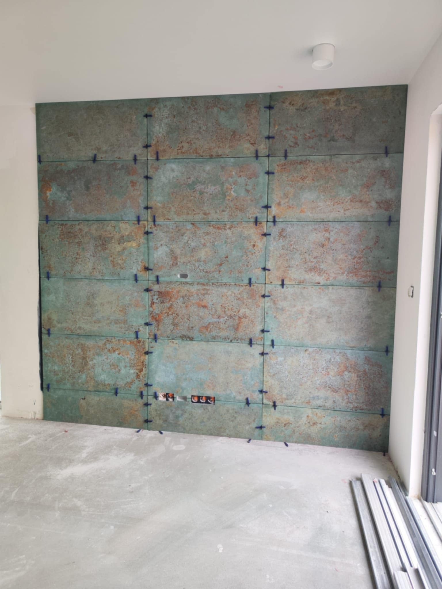 Ściana z płytek imitujących rdzę, w trakcie montażu, z widocznymi krzyżykami dystansowymi, z przygotowanymi otworami na gniazdka elektryczne, na surowym, betonowym podłożu.