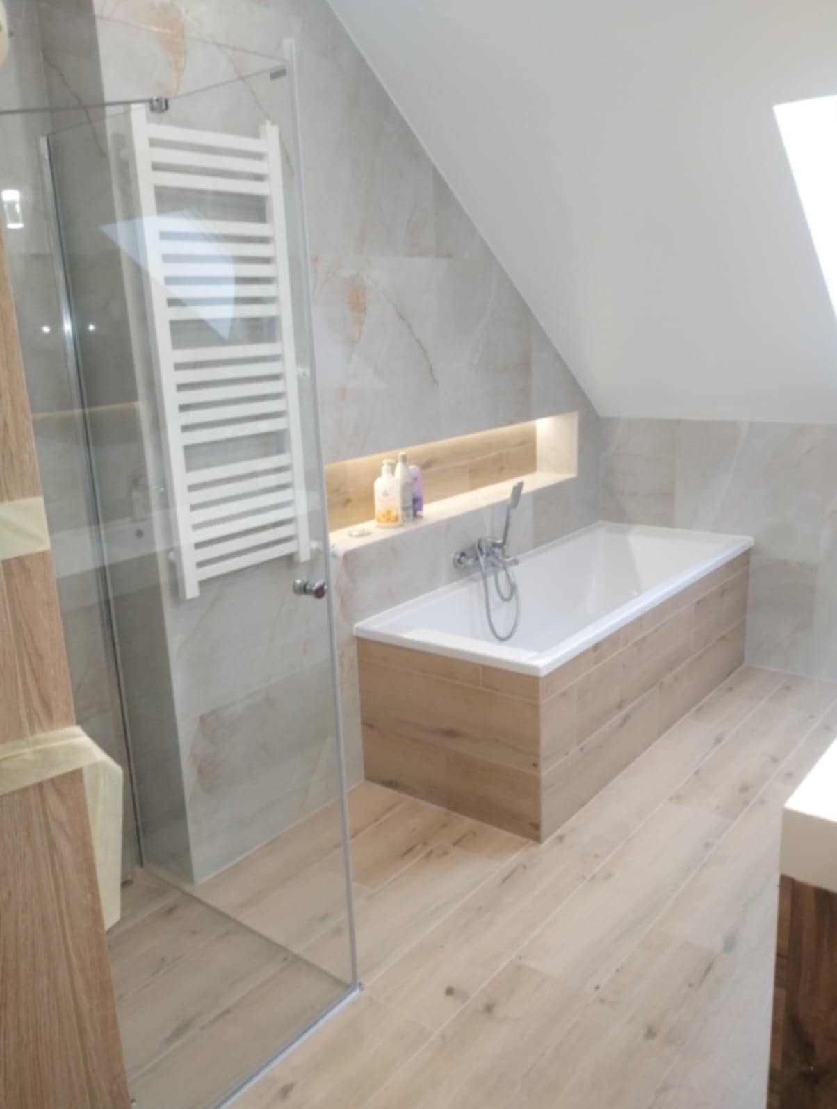 Nowoczesna łazienka na poddaszu z wanną obudowaną panelami imitującymi drewno, przeszkloną kabiną prysznicową i grzejnikiem drabinkowym. Beżowe płytki na ścianach i podłodze, oświetlona wnęka...