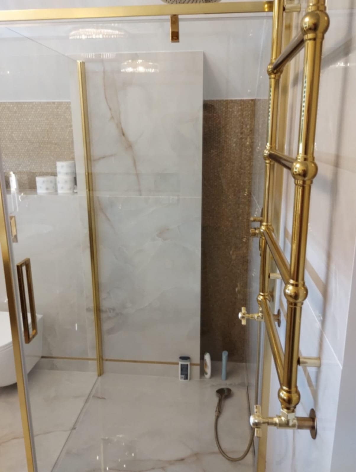 Nowoczesna kabina prysznicowa z elementami w kolorze złotym, widoczny grzejnik łazienkowy w tym samym kolorze, marmurowe płytki na ścianach i podłodze, oraz drobne akcesoria łazienkowe.