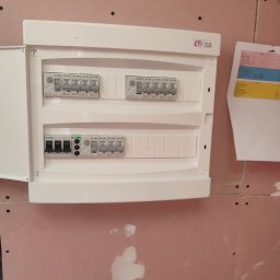 WOJCIECH ANUSZKO EL-WAN - Rozdzielnica elektryczna ETI zamontowana na ścianie z płyt kartonowo-gipsowych, widoczne bezpieczniki i dokumentacja techniczna obok.