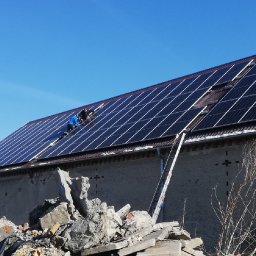 WOJCIECH ANUSZKO EL-WAN - Montaż paneli słonecznych na dachu budynku z gruzem na pierwszym planie. Dwóch monterów w niebieskich kombinezonach pracuje na wysokości, drabina oparta o ścianę.