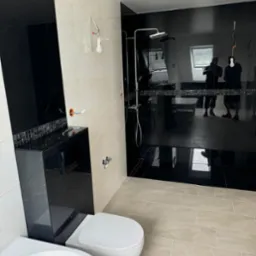 Częściowo wykończona łazienka z czarnymi płytkami imitującymi marmur na ścianie prysznicowej, beżowymi płytkami podłogowymi, i podwieszaną miską WC z czarną obudową; widoczne niedokończone...