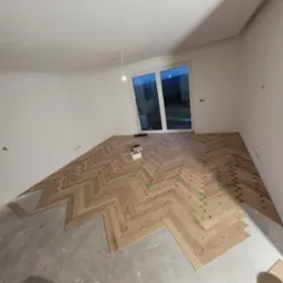 Układanie parkietu w jodełkę, widoczne białe ściany, okno i kliny dystansowe w kolorach czerwonym i zielonym.