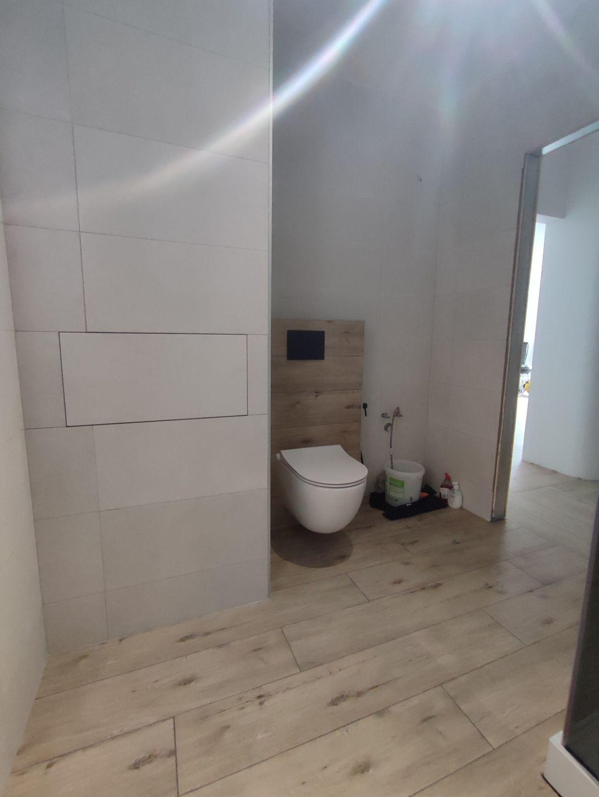 Nowoczesna łazienka z podwieszaną toaletą, drewnopodobną ścianką i jasnymi płytkami podłogowymi, widoczny fragment ściany z ukrytym dostępem rewizyjnym.