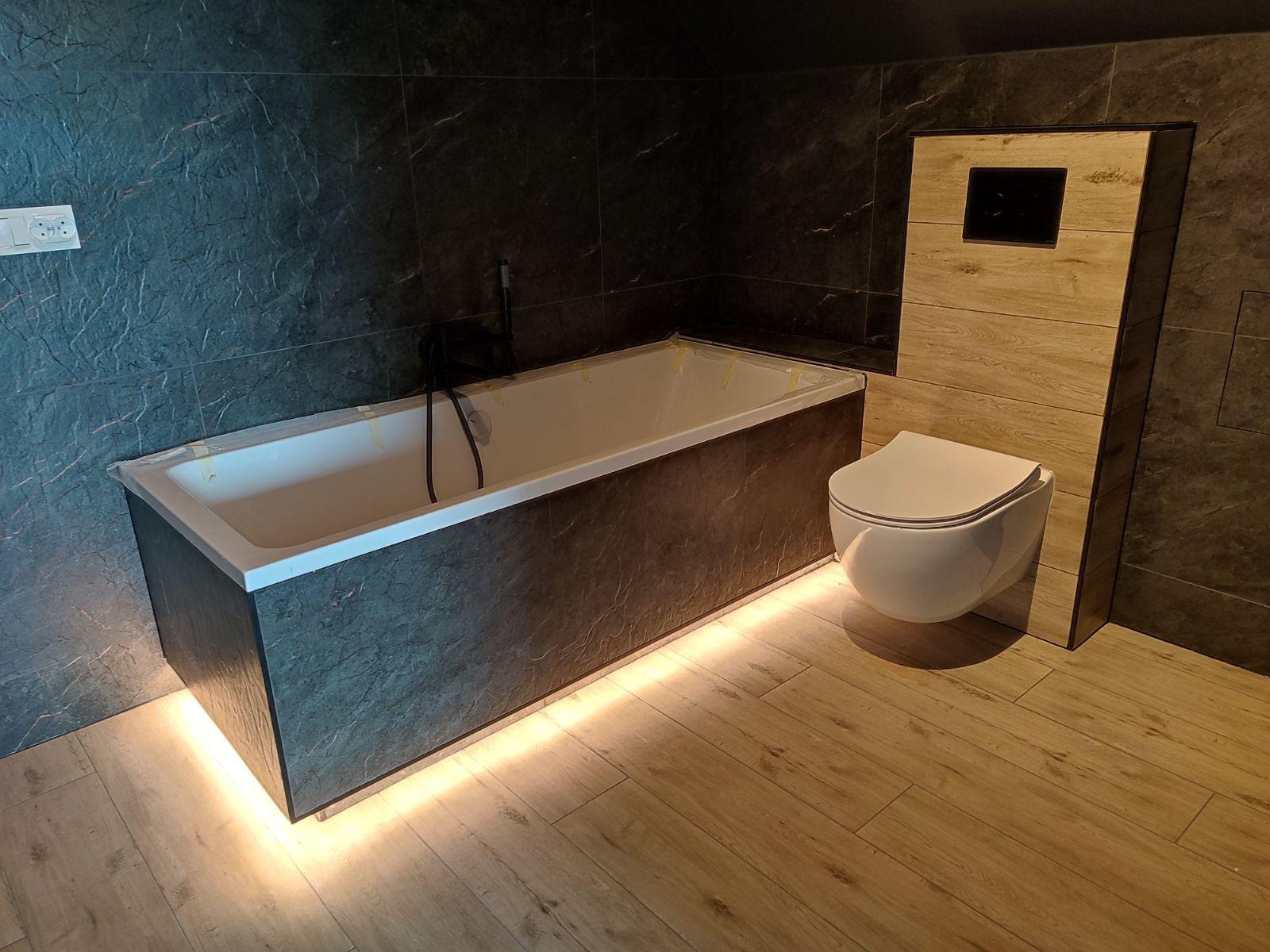 Nowoczesna łazienka z podświetloną wanną obudowaną płytkami imitującymi kamień, podwieszana toaleta z drewnopodobną zabudową, ciemne płytki na ścianach i jasne panele na podłodze.