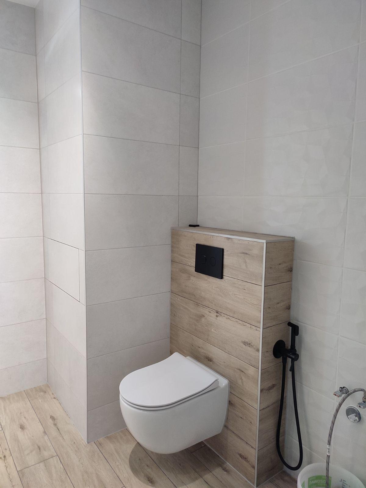 Nowoczesna toaleta podwieszana z drewnianą obudową stelaża, bidetka w kolorze czarnym i jasne płytki w łazience.