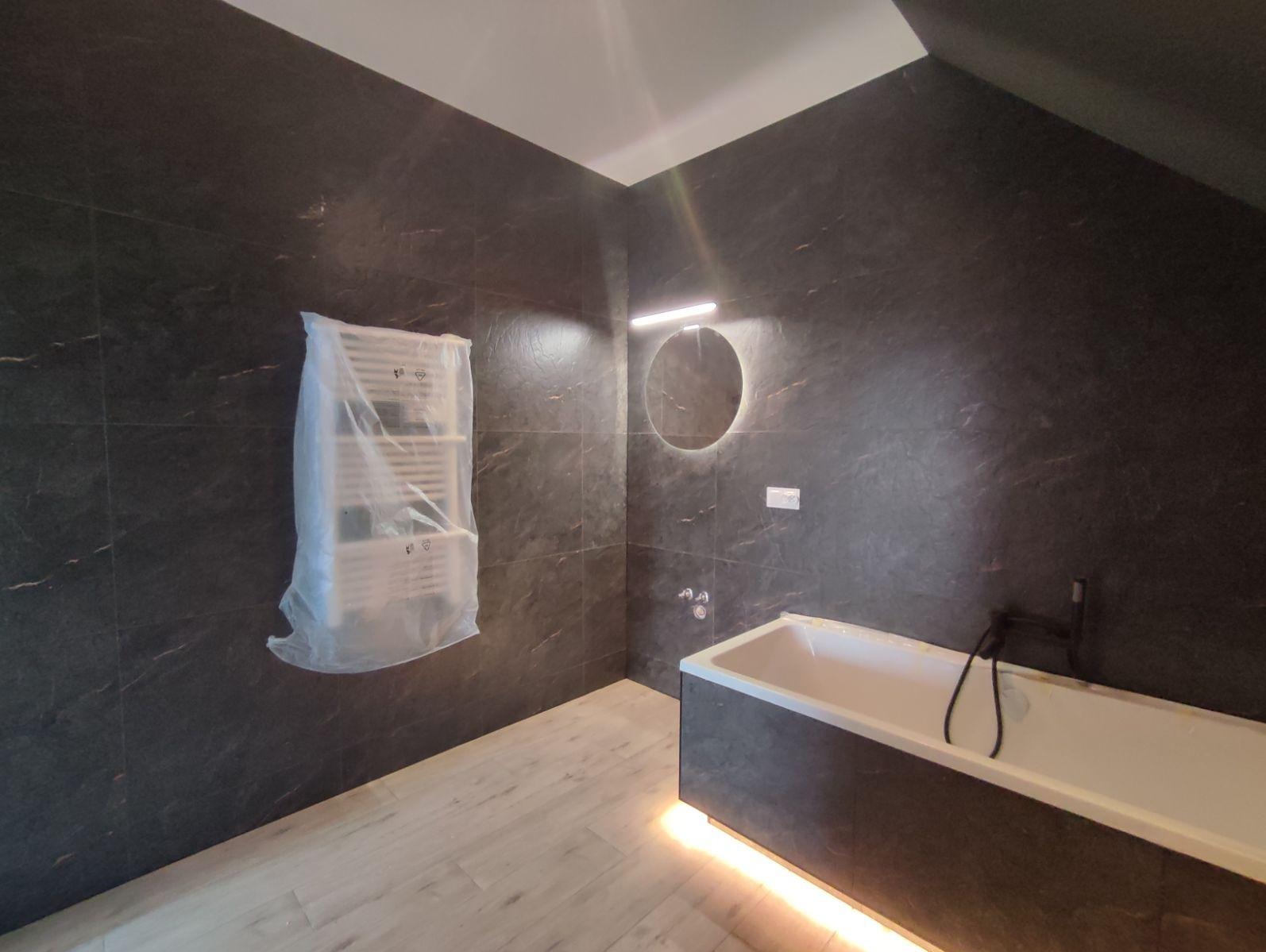 Nowoczesna łazienka w trakcie wykańczania: ciemne płytki imitujące kamień, podświetlana wanna, okrągłe lustro z LED, grzejnik owinięty folią ochronną, jasne panele na podłodze.