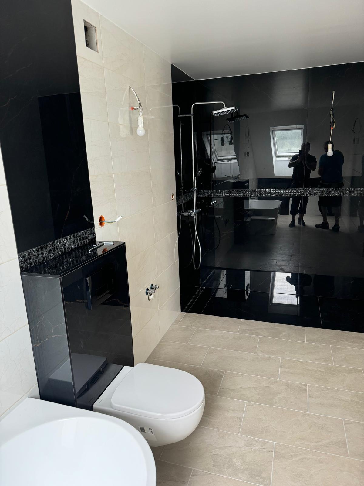 Częściowo wykończona łazienka z czarnymi płytkami imitującymi marmur na ścianie prysznicowej, beżowymi płytkami podłogowymi, i podwieszaną miską WC z czarną obudową; widoczne niedokończone...