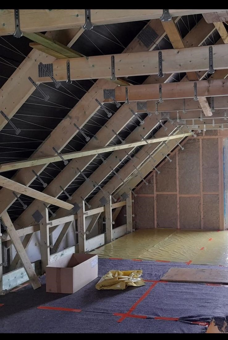 Konstrukcja drewniana poddasza z widocznymi elementami mocującymi, izolacją ścian i folią ochronną na podłodze, karton na pierwszym planie.
