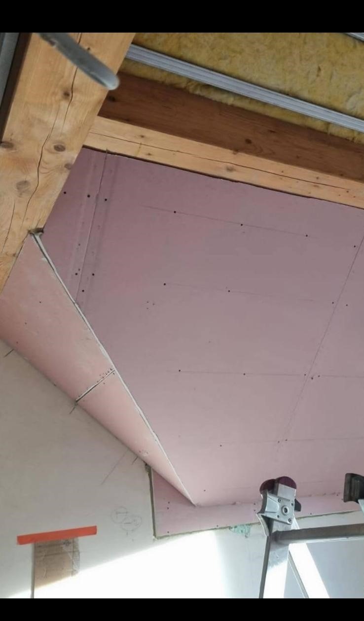 Montaż płyt gipsowo-kartonowych na suficie poddasza, widoczne drewniane belki konstrukcyjne i fragment ocieplenia, metalowa drabina w dolnym rogu.