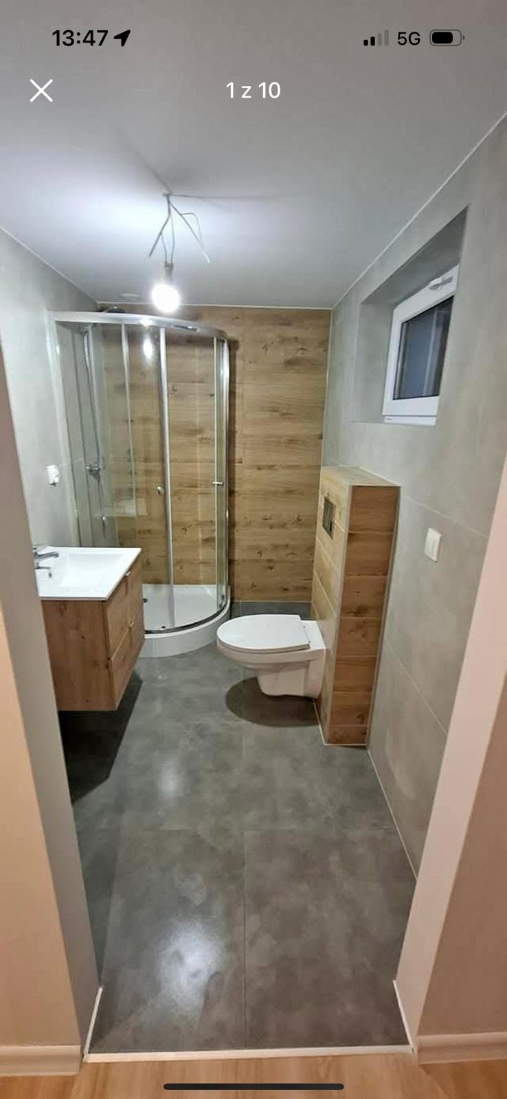 Nowoczesna łazienka z kabiną prysznicową, umywalką z drewnianą szafką, podwieszaną toaletą z obudową z drewnopodobnych paneli oraz szarymi płytkami na ścianach i podłodze. Widoczne niedokończone...
