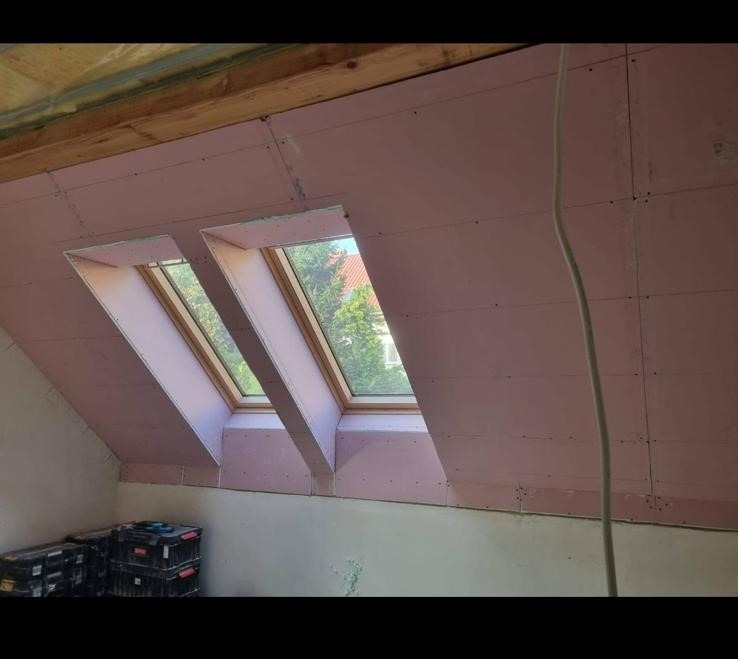 Poddasze z dwoma oknami dachowymi, ściany wyłożone płytami gipsowo-kartonowymi w kolorze różowym, widoczne skrzynki narzędziowe w rogu pomieszczenia.