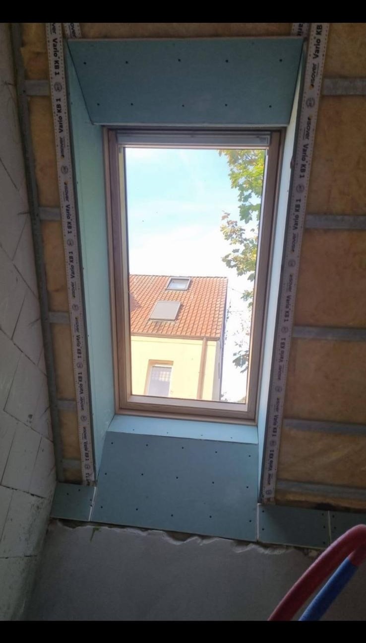 Montaż okna dachowego w trakcie wykańczania poddasza, widoczne płyty gipsowo-kartonowe, izolacja i taśmy uszczelniające, w tle widok na sąsiedni dom z okna.